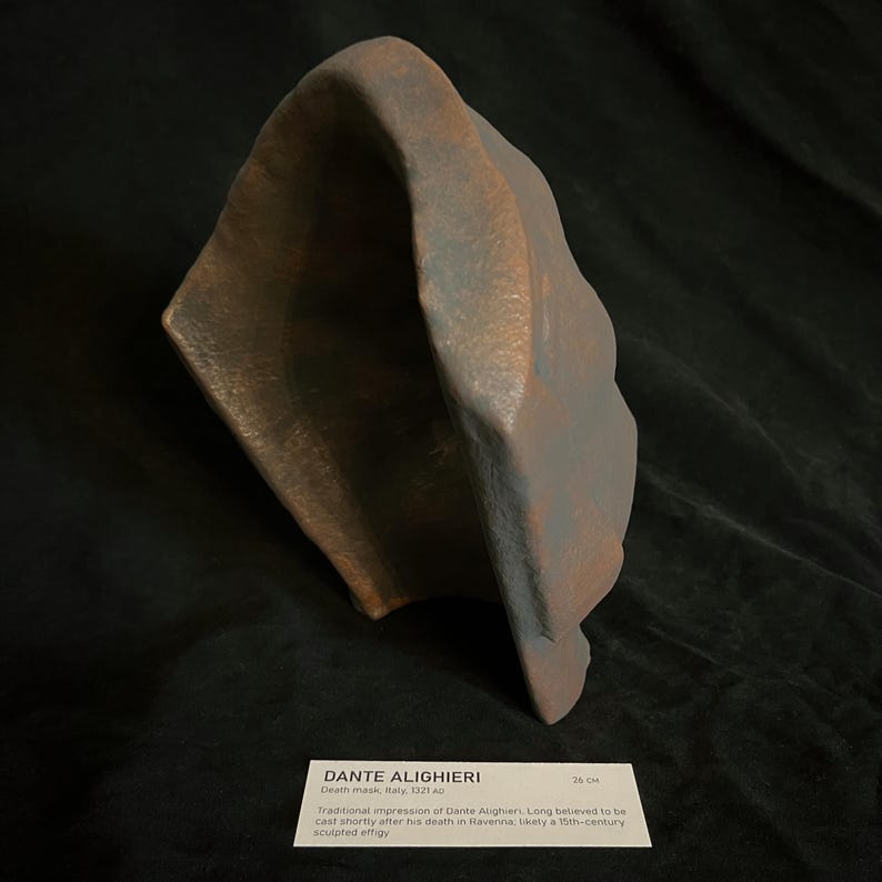 May include: A bronze-colored sculpted effigy of Dante Alighieri, with a hood-like shape. The sculpture is displayed on a dark surface. A label below reads "DANTE ALIGHIERI Death mask, Italy, 1321 AD" and "26 CM".