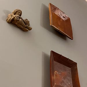 May include: Wall-mounted decorative items. A small, beige sculpture of a figure reading a scroll is mounted on a gray wall. Two square, copper-colored trays are also mounted on the wall, creating a unique display.