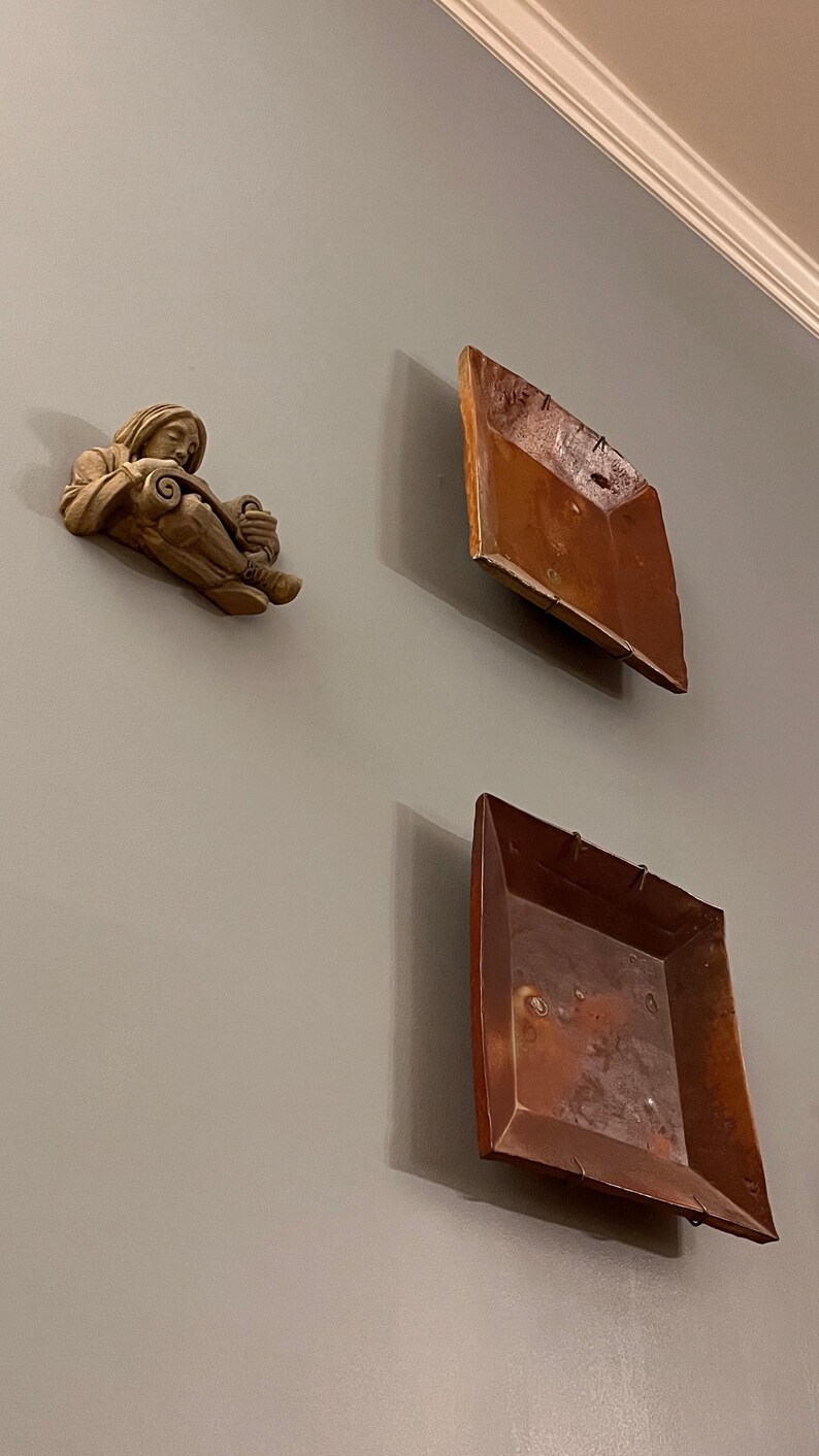 May include: Wall-mounted decorative items. A small, beige sculpture of a figure reading a scroll is mounted on a gray wall. Two square, copper-colored trays are also mounted on the wall, creating a unique display.