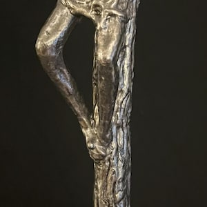 May include: A detailed sculpture of a human figure, crafted from a metallic material. The artwork depicts the lower portion of the body, with a leg bent at the knee and a textured tree trunk-like form. The sculpture has a dark, aged appearance.