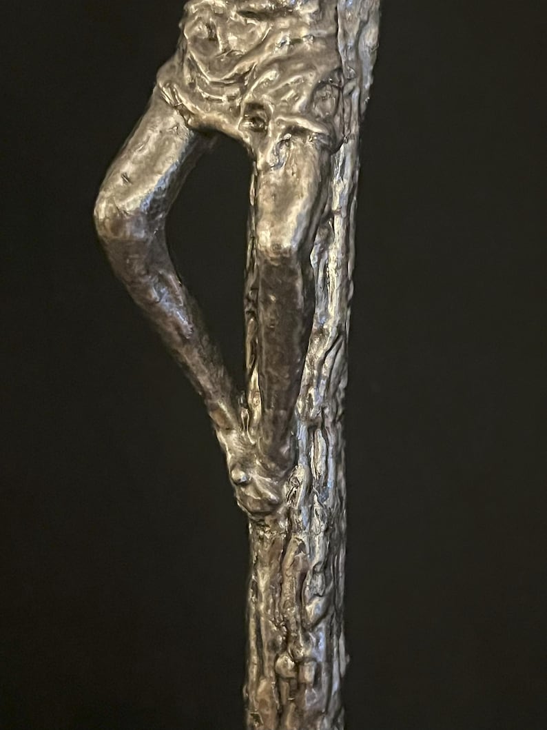 May include: A detailed sculpture of a human figure, crafted from a metallic material. The artwork depicts the lower portion of the body, with a leg bent at the knee and a textured tree trunk-like form. The sculpture has a dark, aged appearance.