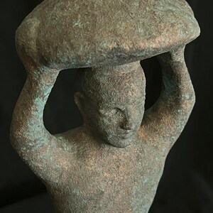 May include: A bronze sculpture of a person carrying a large, round object on their head. The sculpture has a weathered, green patina.