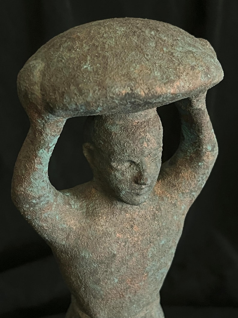 May include: A bronze sculpture of a person carrying a large, round object on their head. The sculpture has a weathered, green patina.