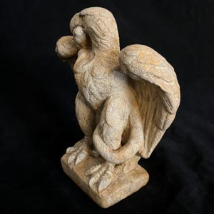 May include: A weathered, beige-colored griffin statue. The mythical creature has a bird-like head, large wings, and the body of a lion. The statue is perched on a square base, showcasing intricate details and textures.
