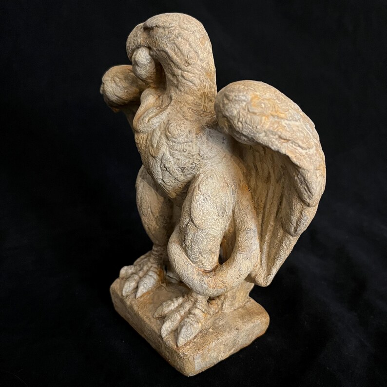 May include: A weathered, beige-colored griffin statue. The mythical creature has a bird-like head, large wings, and the body of a lion. The statue is perched on a square base, showcasing intricate details and textures.