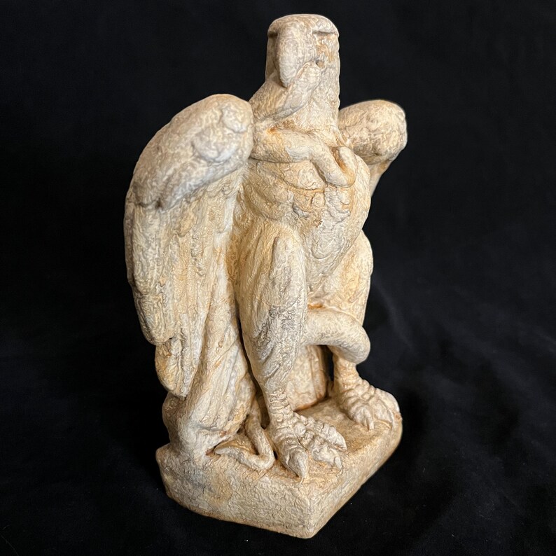 May include: A beige-colored, stone-like sculpture of a griffin, a mythical creature with the body of a lion and the head and wings of an eagle. The sculpture is detailed with textured wings, feathers, and claws, and stands on a square base.