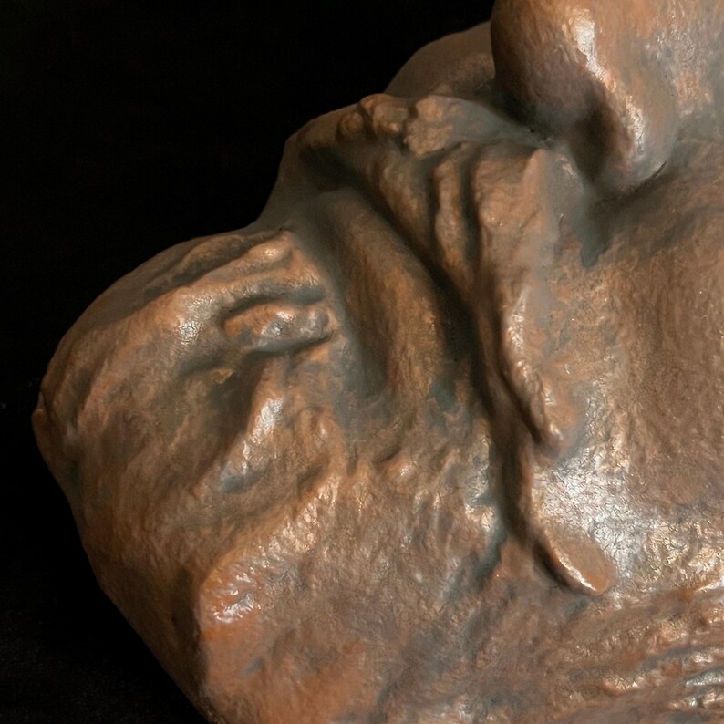 Puede incluir: Primer plano de una escultura de color bronce, posiblemente un busto o relieve. La obra de arte tiene una superficie texturizada con detalles visibles, incluyendo pliegues y contornos. La iluminaci&oacute;n resalta los tonos marrones y el dise&ntilde;o intrincado.