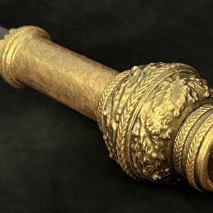 May include: Close-up of a decorative staff or sceptre. The staff features a dark handle with an ornate gold-coloured metalwork design. The metalwork includes intricate patterns and a circular end piece.