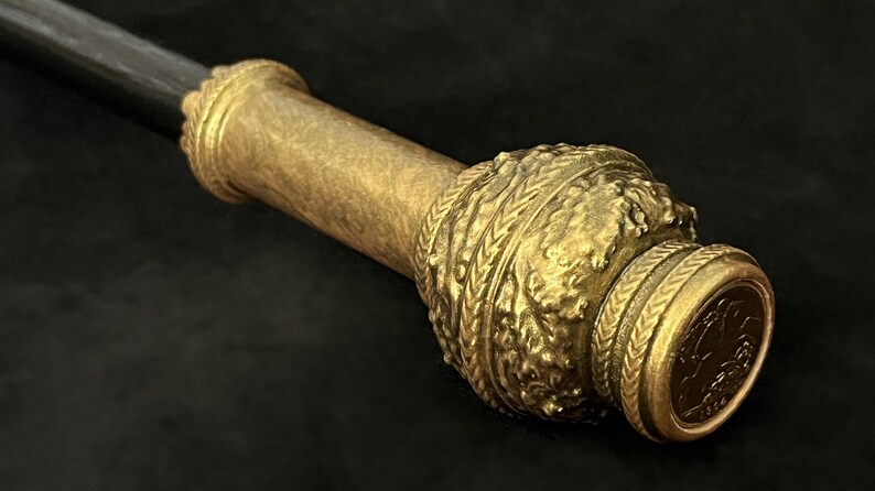 May include: Close-up of a decorative staff or sceptre. The staff features a dark handle with an ornate gold-coloured metalwork design. The metalwork includes intricate patterns and a circular end piece.