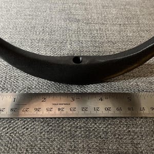 May include: Black metal hook with a curved shape, measuring approximately 10 cm in length. The hook is shown against a grey fabric background.