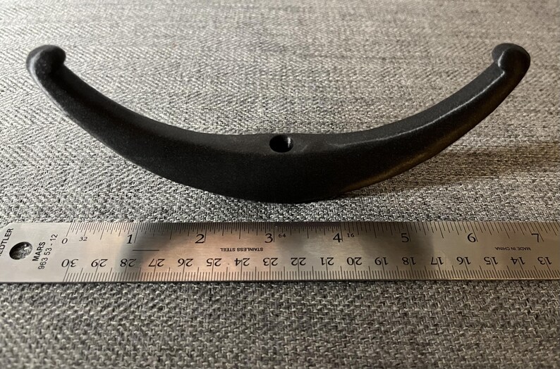May include: Black metal hook with a curved shape, measuring approximately 10 cm in length. The hook is shown against a grey fabric background.