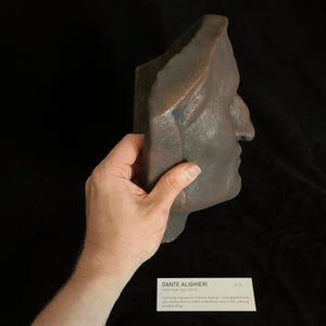 May include: A bronze-colored sculpture of a side profile of a face, held by a hand. The sculpture is of Dante Alighieri, a traditional expression of Dante Alighieri. The sculpture is on a black background.