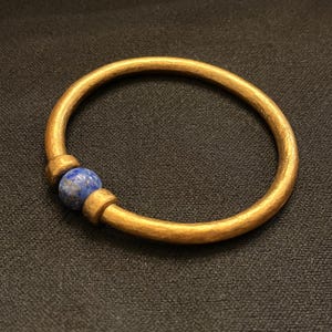 May include: A gold-coloured bracelet featuring a blue lapis lazuli bead. The circular bracelet is made of metal. The bead is positioned centrally, providing a contrast. The bracelet is presented on a dark surface. The bracelet's diameter is approximately 6.5 cm.