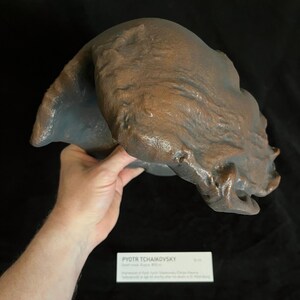 Puede incluir: Una m&aacute;scara mortuoria de color bronce de Piotr Chaikovski, con una superficie texturizada y una representaci&oacute;n detallada de los rasgos faciales. La m&aacute;scara est&aacute; sostenida por una mano, y una peque&ntilde;a tarjeta informativa es visible debajo.