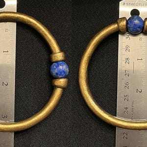 May include: Two gold-coloured bangles with a blue stone accent. The bangles are circular and appear to be made of metal. A ruler is placed next to each bangle, showing the size of the jewellery.