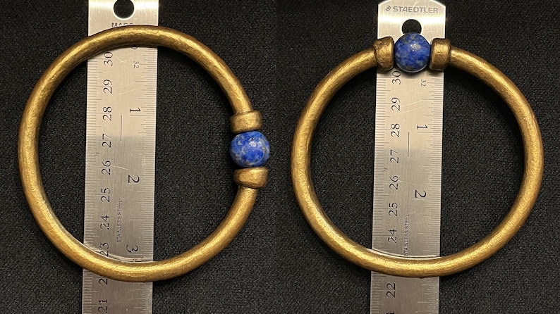 May include: Two gold-coloured bangles with a blue stone accent. The bangles are circular and appear to be made of metal. A ruler is placed next to each bangle, showing the size of the jewellery.