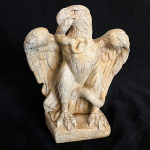 May include: A detailed, off-white eagle statue with outstretched wings and a thoughtful pose. The bird's talons are visible, and the statue is on a square base. The sculpture has a textured surface and is likely made of stone or a similar material.
