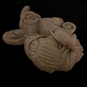 May include: A detailed, beige-colored sculpture with a complex, organic design. The sculpture features a combination of rounded and swirling shapes, with a textured surface. The piece appears to be made of stone or a similar material, and is set against a black background.