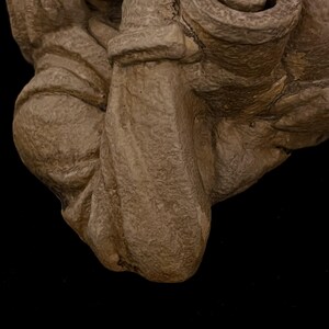 May include: Close-up of a beige, textured sculpture with a rough, aged appearance. The sculpture has rounded shapes and folds, suggesting a draped or organic form. The background is solid black, highlighting the sculpture's details.