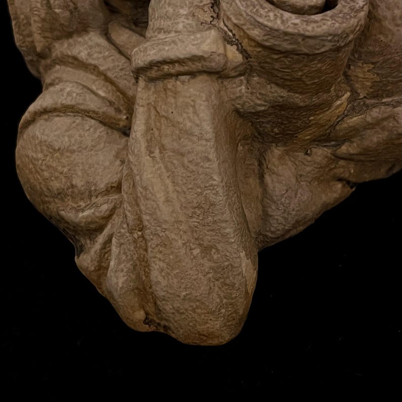 May include: Close-up of a beige, textured sculpture with a rough, aged appearance. The sculpture has rounded shapes and folds, suggesting a draped or organic form. The background is solid black, highlighting the sculpture's details.