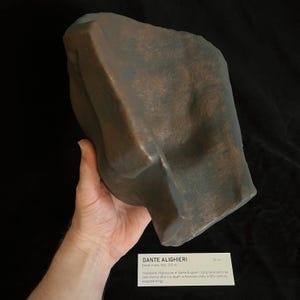 May include: A bronze-colored death mask of Dante Alighieri, with a textured surface. The mask is held in a hand. Text on a card reads "DANTE ALIGHIERI Death mask, Italy, 1321". The mask is approximately 10 inches in length.
