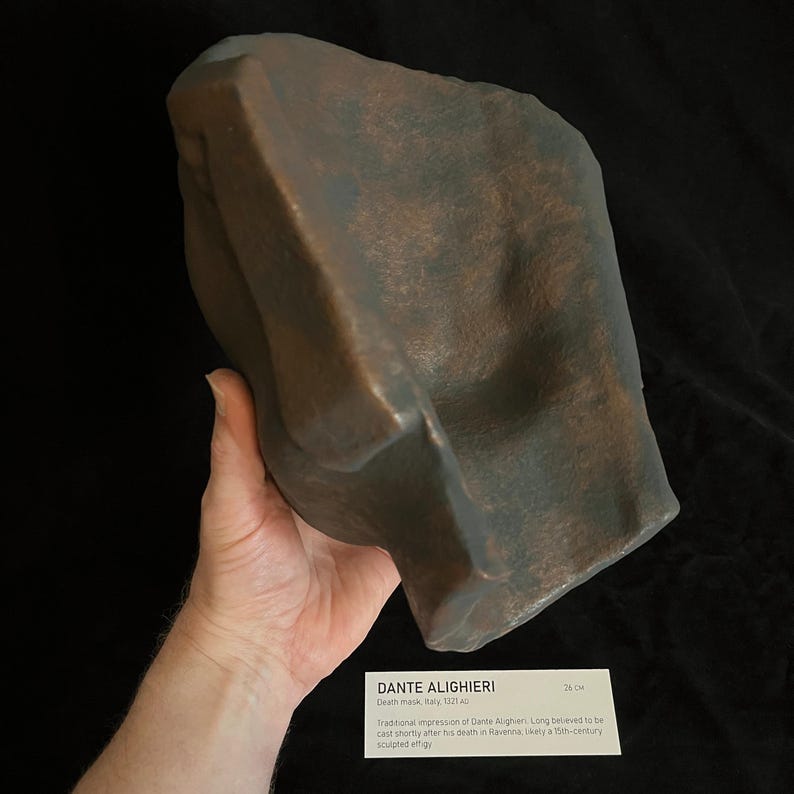 May include: A bronze-colored death mask of Dante Alighieri, with a textured surface. The mask is held in a hand. Text on a card reads "DANTE ALIGHIERI Death mask, Italy, 1321". The mask is approximately 10 inches in length.
