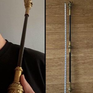 May include: A decorative wand with a gold-coloured lion head and ornate detailing. The wand is black with gold accents and measures approximately 104 cm in length. The wand is held by a person wearing a black shirt.
