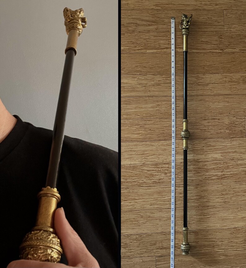 May include: A decorative wand with a gold-coloured lion head and ornate detailing. The wand is black with gold accents and measures approximately 104 cm in length. The wand is held by a person wearing a black shirt.
