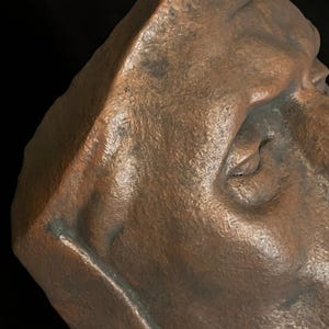 May include: Close-up of a bronze-colored sculpture of a human face, featuring detailed textures and shadows. The sculpture's surface has a weathered appearance, with visible imperfections and subtle color variations.