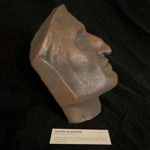 May include: A bronze-colored sculpted effigy of Dante Alighieri, a profile view with a prominent nose and hooded garment. The image includes a label with the text "DANTE ALIGHIERI" and details about the death mask, including the measurement of 76 cm.
