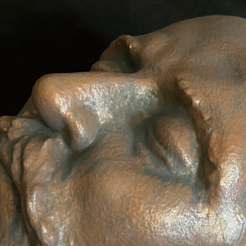 Puede incluir: Primer plano de una escultura de color bronce de un rostro humano de perfil. La escultura tiene una superficie texturizada y muestra el ojo, la nariz y parte de la boca. El fondo es oscuro.