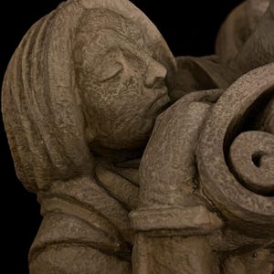 May include: A close-up of a stone sculpture depicting a person with closed eyes, resting their head against a decorative, swirling element. The sculpture is a muted gray color, with visible texture and detail.