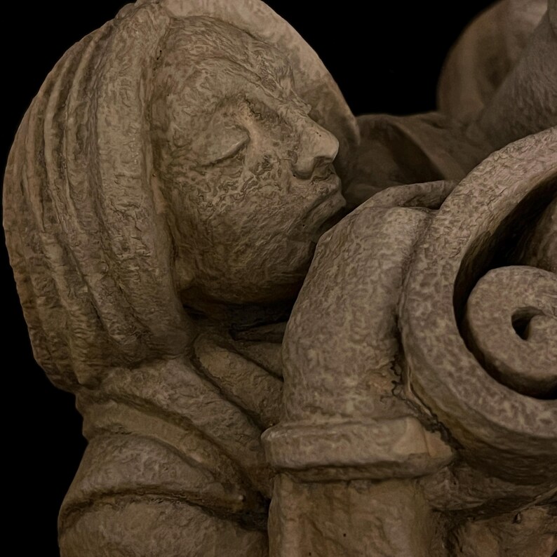 May include: A close-up of a stone sculpture depicting a person with closed eyes, resting their head against a decorative, swirling element. The sculpture is a muted gray color, with visible texture and detail.