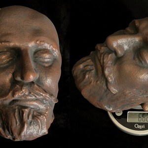 Puede incluir: Una escultura de color bronce de la cara de un hombre con los ojos cerrados y una barba completa. La escultura se muestra en dos vistas, una frontal y otra de perfil, con una regla para la escala. La vista de perfil descansa sobre una b&aacute;scula, con un peso de 188 gramos.