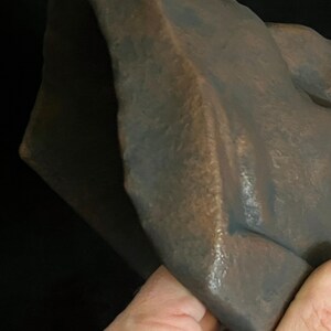 May include: A close-up of a bronze-colored sculpture with a textured surface. The sculpture has a folded, draped appearance, with visible ridges and shadows. The artwork is held by a hand.