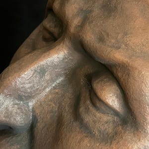 May include: Close-up of a bronze-colored sculpture of a human face. The sculpture has detailed features, including a nose, eye, and cheek, with visible texture and shadows. The background is dark.