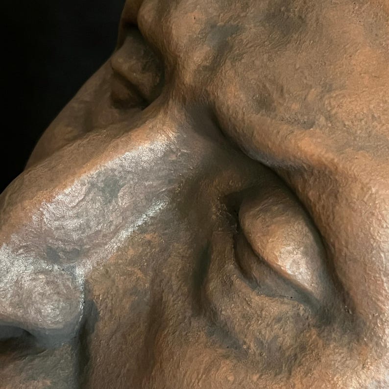 May include: Close-up of a bronze-colored sculpture of a human face. The sculpture has detailed features, including a nose, eye, and cheek, with visible texture and shadows. The background is dark.