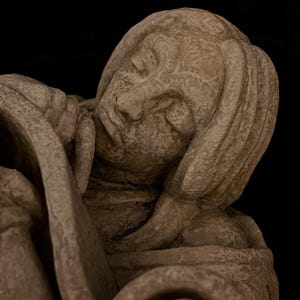 May include: A detailed sculpture of a person with their head resting on their arm. The sculpture is a light beige color and has a textured surface. The person's face is visible, with closed eyes and a serene expression. The background is black.