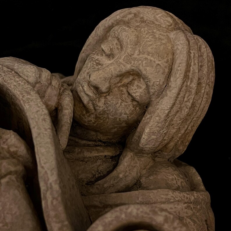 May include: A detailed sculpture of a person with their head resting on their arm. The sculpture is a light beige color and has a textured surface. The person's face is visible, with closed eyes and a serene expression. The background is black.