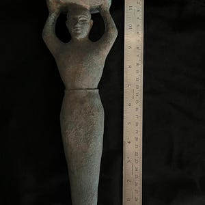 May include: A green bronze sculpture of a person carrying a large object on their head. The sculpture is about 11 inches tall and is made in a stylized, simplified form.
