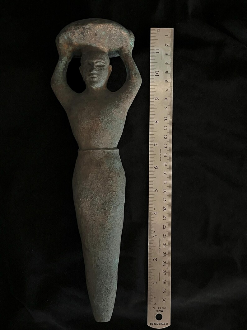 May include: A green bronze sculpture of a person carrying a large object on their head. The sculpture is about 11 inches tall and is made in a stylized, simplified form.