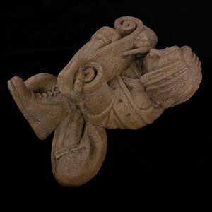 May include: A detailed, brown sculpture of a person in a seated position, holding a scroll. The figure is wearing a hat and boots. The sculpture has a textured surface and is set against a black background.