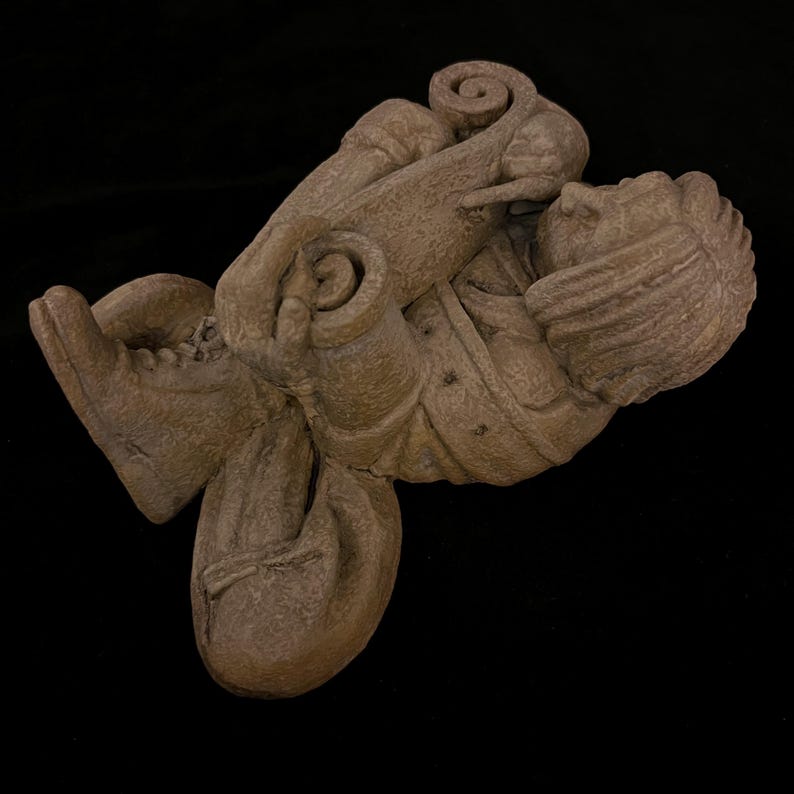 May include: A detailed, brown sculpture of a person in a seated position, holding a scroll. The figure is wearing a hat and boots. The sculpture has a textured surface and is set against a black background.