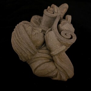 May include: A detailed, brown sculpture of a person holding a scroll and a container. The sculpture has a textured surface and is set against a black background. The figure's face is visible, and the scroll has a rolled design.