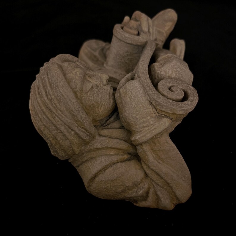 May include: A detailed, brown sculpture of a person holding a scroll and a container. The sculpture has a textured surface and is set against a black background. The figure's face is visible, and the scroll has a rolled design.