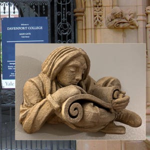 May include: A beige stone sculpture of a person reading a scroll. The sculpture is mounted on a wall. In the background is a sign that reads "Welcome to Davenport College" and "Main Gate York Street."