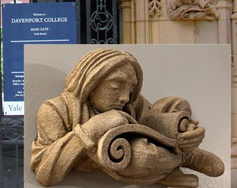 May include: A beige stone sculpture of a person reading a scroll. The sculpture is mounted on a wall. In the background is a sign that reads "Welcome to Davenport College" and "Main Gate York Street."