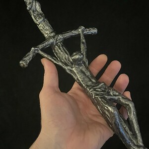 May include: A detailed, metallic sculpture of a figure on a cross. The sculpture is held in a hand, showcasing the intricate details of the figure and the cross. The piece has a textured, aged appearance.
