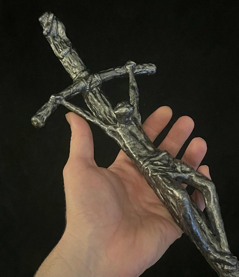 May include: A detailed, metallic sculpture of a figure on a cross. The sculpture is held in a hand, showcasing the intricate details of the figure and the cross. The piece has a textured, aged appearance.