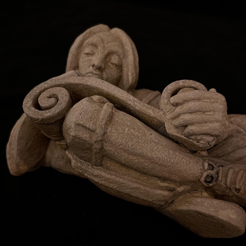 May include: A detailed sculpture of a person in a reclining pose, crafted from a light brown material. The figure holds a scroll, with closed eyes, suggesting a moment of contemplation. The sculpture's texture and form are highlighted against a black background.